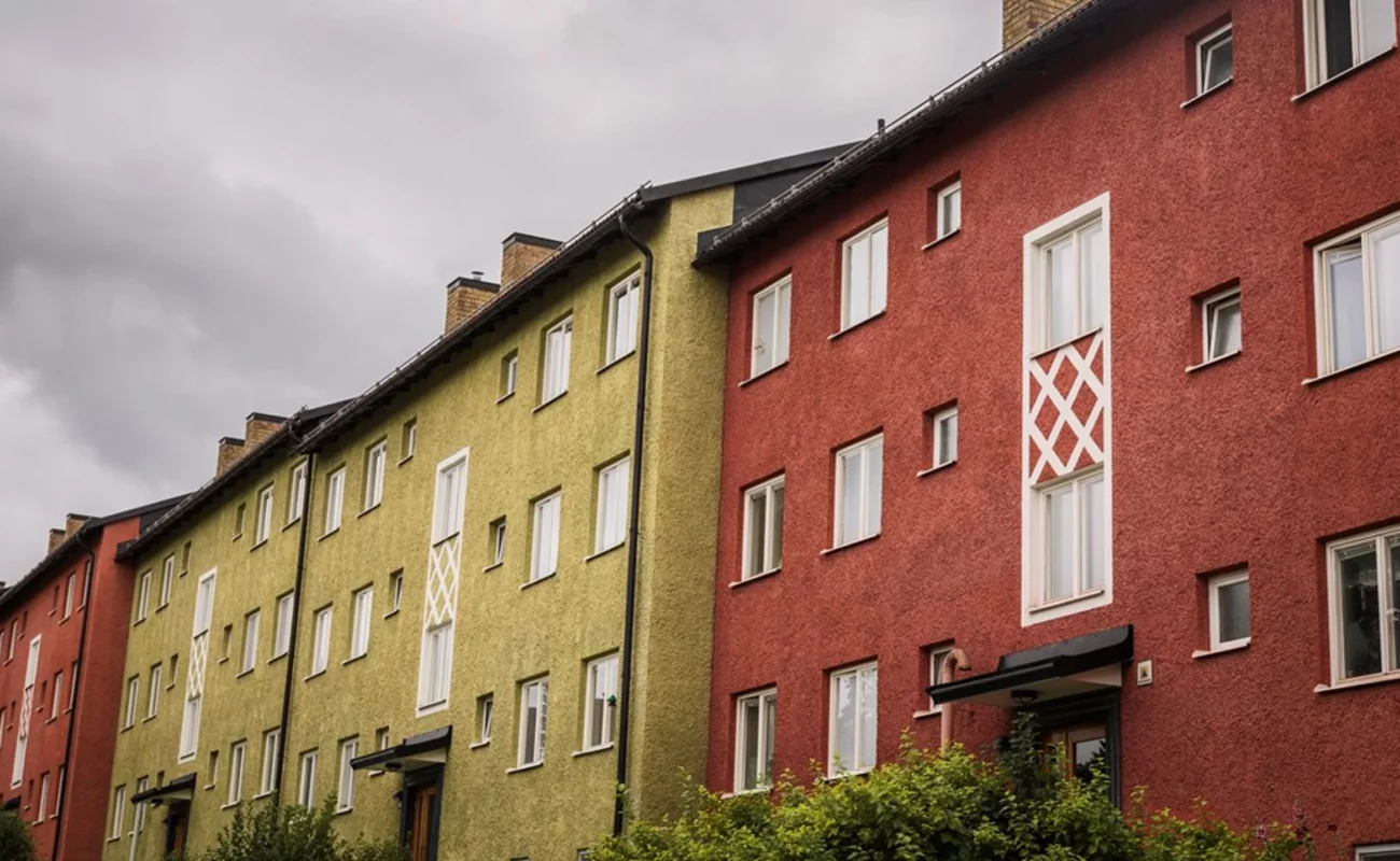 En rad färgglada lägenhetsbyggnader med en texturerad putsad fasad i rött och grönt. Byggnaderna har stora vita dekorativa fönsterramar och små fönster. Himlen är mulen med mörka moln som ger en dramatisk stämning.