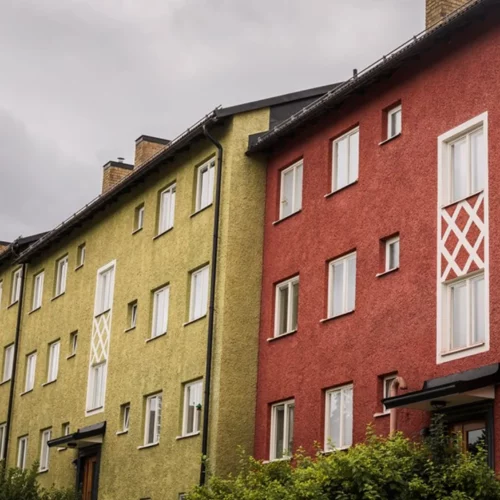 En rad färgglada lägenhetsbyggnader med en texturerad putsad fasad i rött och grönt. Byggnaderna har stora vita dekorativa fönsterramar och små fönster. Himlen är mulen med mörka moln som ger en dramatisk stämning.